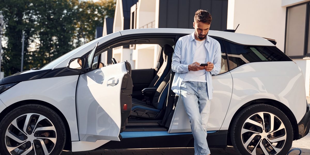 Person står lutad mot en vit elbil med öppna dörrar och tittar på en mobiltelefon. Bilen är parkerad på en uppfart med moderna hus i bakgrunden.