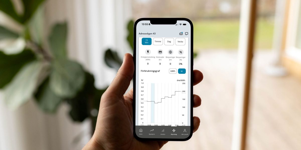 En hand håller en smartphone som visar Kalmar Energis app med realtidsdata för energianvändning. Skärmen visar en graf över förbrukning i öre per kWh och olika menyval för tidsintervall.