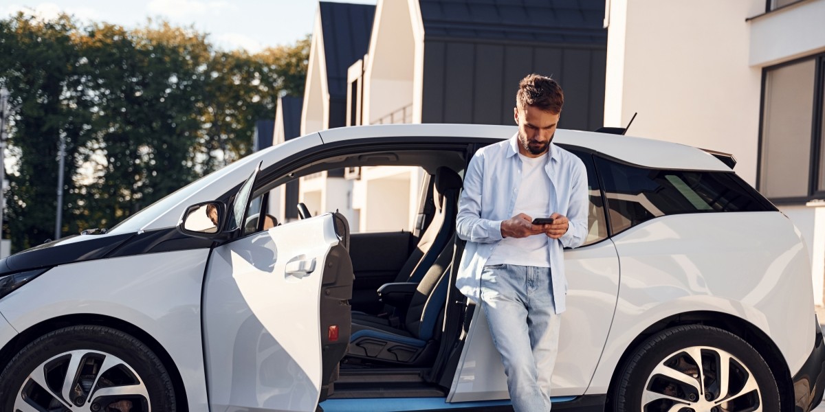 Person står lutad mot en vit elbil med öppna dörrar och tittar på en mobiltelefon. Bilen är parkerad på en uppfart med moderna hus i bakgrunden.