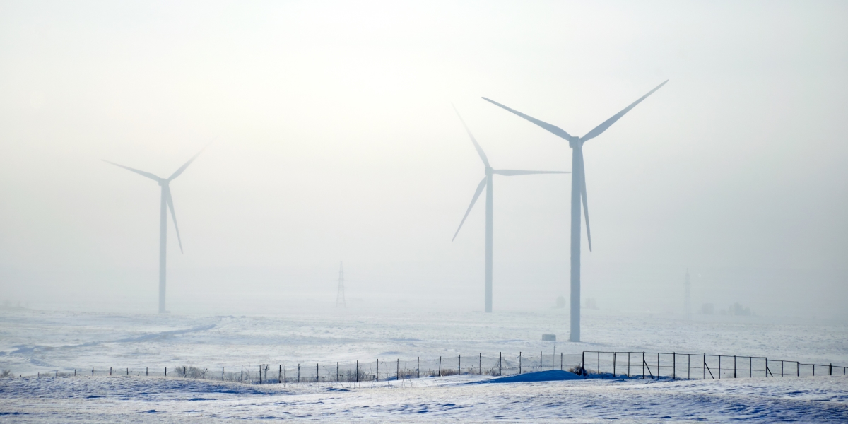 Tre vindkraftverk står i ett snötäckt landskap under dimmiga vinterförhållanden. Sikten är begränsad och marken är täckt av frost och snö, med ett stängsel i förgrunden.