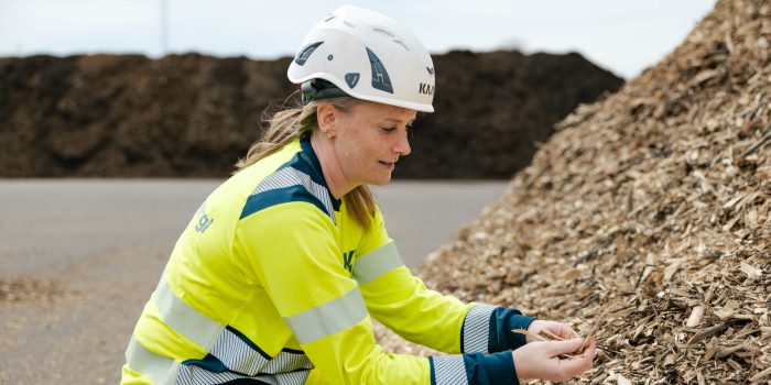 Medarbetare på Kalmar Energi i varselkläder och skyddshjälm sitter vid en stor hög med träflis och håller flisbitar i händerna utomhus på Moskogen.